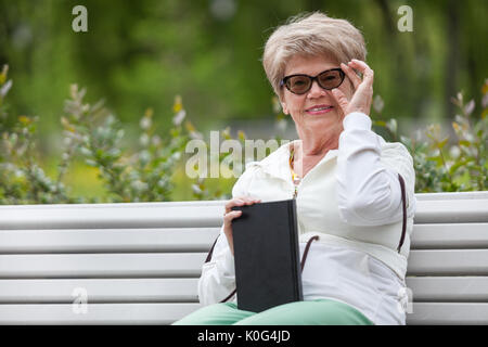 Senior sorridente donna che guarda la fotocamera, tenendo gli occhiali e libro nero, d'estate il parco Foto Stock