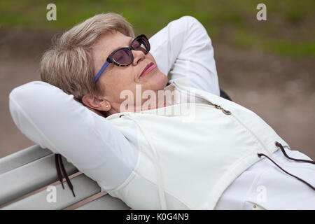 Un anziano donna in bicchieri di dormire sulla sua schiena sul banco in estate park Foto Stock