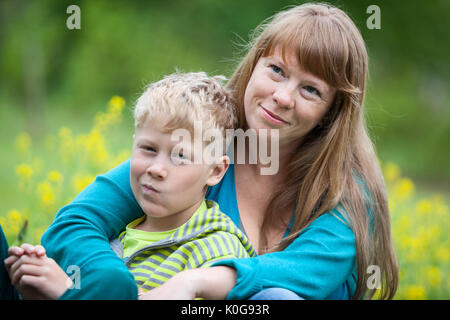 Mamma e Bambino sono seduti su un prato estivo tra i fiori. Donna che abbraccia il suo figlio Foto Stock