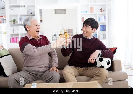 Sorridente uomo senior e il suo giovane figlio tostare con bottiglie di birra guardando la partita di calcio in TV Foto Stock