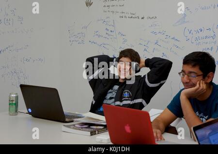 Due studenti maschi seduti a un tavolo bianco con due computer portatili, materiali di studio e una bevanda energetica in scatola; pareti di lavagna bianca contenenti scrittura ed equazioni in entrambi i marcatori blu e nero, espressioni facciali sorridenti, 2016. Cortesia Eric Chen. Foto Stock