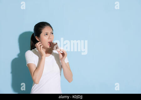 Attraente bella signora in piedi in blu sullo sfondo della parete cercando di mangiare il cioccolato dolce cibo e guardando all'area vuota preoccupati per le cavità dentali p Foto Stock