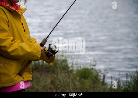 Giovane ragazza in un impermeabile giallo pesca accanto a un lago Foto Stock