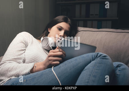 Ragazza rilassante sul divano di casa a tarda notte, ella è il collegamento con una tavoletta digitale e di indossare le cuffie Foto Stock