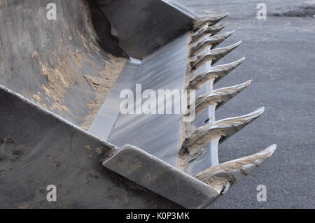Svuotare la benna di un escavatore con tracce di sabbia e terra sulla carta lucida i denti e le superfici contro lo sfondo di asfalto. Foto Stock