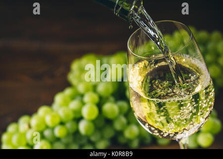 Versare il vino bianco in un bicchiere con un grappolo di uva verde contro lo sfondo di legno Foto Stock