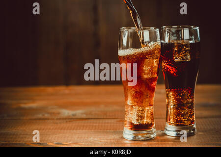 Versando bella bevanda fredda di cola con cubetti di ghiaccio con una caldaia cannucce in bicchieri su sfondo di legno con spazio libero Foto Stock