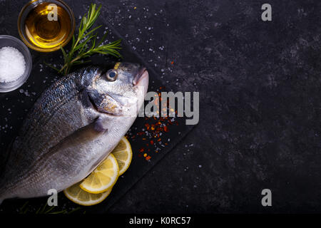 Crudo fresco Orate di mare il pesce con le fette di limone, sale, rosmarino e olio d'oliva sul nero ardesia sfondo. Cibo sano concetto, vista dall'alto, spazio di copia Foto Stock