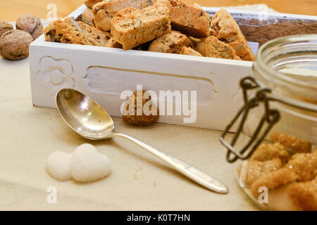 Italian tozzetti biscotti in un bianco scatola di legno con un cucchiaio d'argento e a forma di cuore i cubetti di zucchero. Formato orizzontale. Foto Stock
