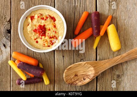 Baby rainbow carote con hummus dip e cucchiaio, vista aerea su un rustico sfondo di legno Foto Stock