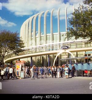 Ammira l'esterno della mostra Ford in un pomeriggio pieno di impegni alla fiera del mondo di New York, a Flushing Meadows, Queens, New York City, giugno 1964. I visitatori del parco camminano davanti a una grande banca di telefoni pubblici. Un gianitore afroamericano spazza la lettiera al telaio centrale. Sullo sfondo, è visibile il giro in Magic Skyway di Ford, con i carabieri seduti in auto convertibili che corrono su un tracciato all'interno della mostra di vetro in padiglione. Foto Stock