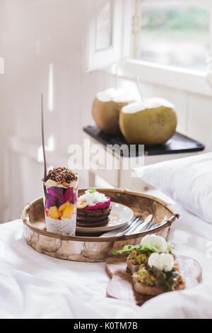 Fresh gustosa colazione a letto su vassoio in legno Yogurt con frutta e chia, frittelle di miele, due toast con avocado e uova in camicia e due giovani cocon Foto Stock