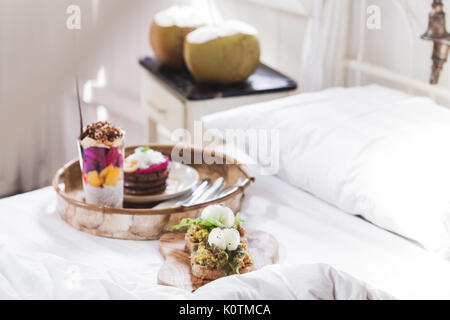 Fresh gustosa colazione a letto su vassoio in legno Yogurt con frutta e chia, frittelle di miele, due toast con avocado e uova in camicia e due giovani cocon Foto Stock