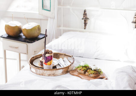Fresh gustosa colazione a letto su vassoio in legno Yogurt con frutta e chia, frittelle di miele, due toast con avocado e uova in camicia e due giovani cocon Foto Stock