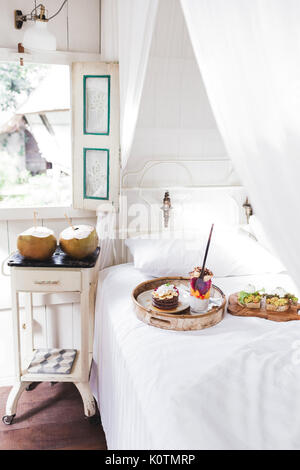 Fresh gustosa colazione a letto su vassoio in legno Yogurt con frutta e chia, frittelle di miele, due toast con avocado e uova in camicia e due giovani cocon Foto Stock