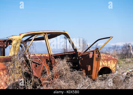 Abbandonato e arrugginito relitto di un giallo sovietica vintage vettura russa nel mezzo di fieno secco con scenic ghiaccio montagne superiore nel sud dell'Armenia. Foto Stock