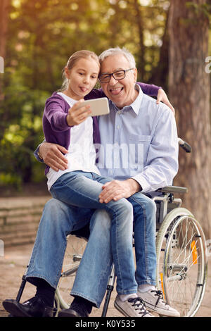 Funny nipote usando il telefono per selfie con suo padre disabili in sedia a rotelle e madre nel parco Foto Stock