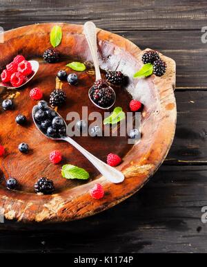 Fresche bacche mature lamponi, more e mirtilli su un marrone piatto di legno con un cucchiaio su tavoli in legno nero, rustico, il fuoco selettivo, spa Foto Stock