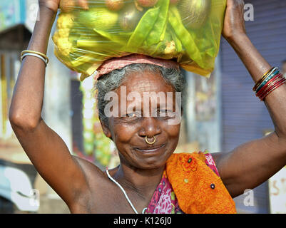 La donna indiana anziana Adivasi (Orissan tribal woman) con un anello dorato del naso trasporta sulla sua testa un sacchetto pesante di plastica pieno di verdure assortite. Foto Stock