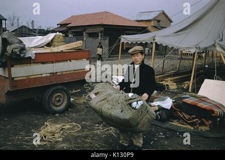 Uomo giapponese in un villaggio che porta un sacco di tessili, avvolti in un fascio, carrello visibile alla sua sinistra, pile di coperte sul terreno e un partito tenda parzialmente assemblati in background, Giappone, 1952. Foto Stock
