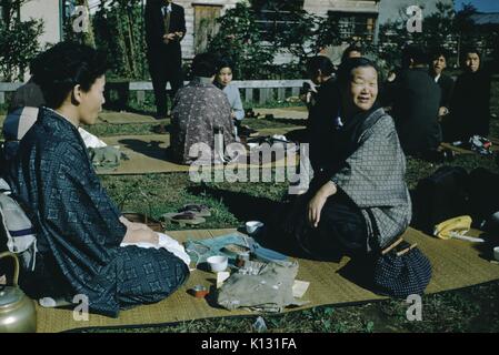 Due donne giapponesi inginocchiati su una stuoia di paglia, affacciati e godendo di un picnic nel cortile della chiesa ad una chiesa della missione, altri gruppi su stuoie visibile in background, Giappone, 1952. Foto Stock