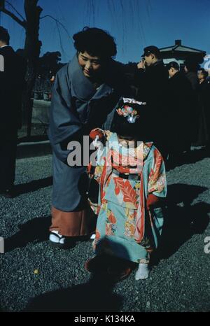 Donna giapponese la piegatura verso il basso per tenere la mano di sua figlia, che indossa un rosso e blu kimono, una folla di uomini in background in piedi al di fuori di un tempio, Giappone, 1951. Foto Stock