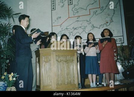 Il coro delle ragazze giapponesi, con il predicatore, cantare dai libri di inni cristiani in una Chiesa missionaria, una mappa del Giappone sulla parete in backround, Giappone, 1951. Foto Stock