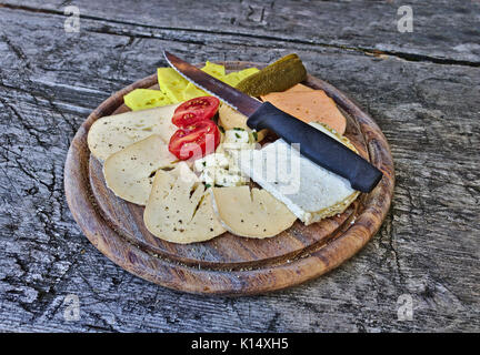 Vari formaggi diversi disposti su un piatto di legno con pomodori, sottaceti e il coltello. Foto Stock