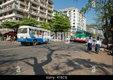 Due uomini birmani a piedi lungo una strada trafficata in Yangon Myanmar con edifici moderni e gli autobus dietro di loro Foto Stock