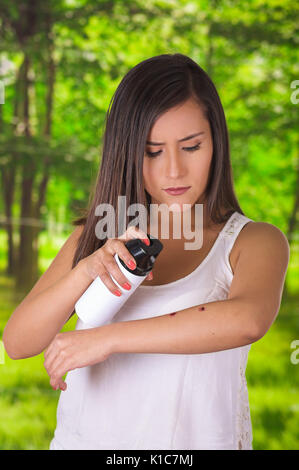 Ravvicinata di una giovane donna che soffre di prurito dopo punture di zanzara, utilizzando una spruzzatrice di oltre il morso di insetto, allergiche della pelle il concetto di trattamento, in una foresta verde sullo sfondo Foto Stock