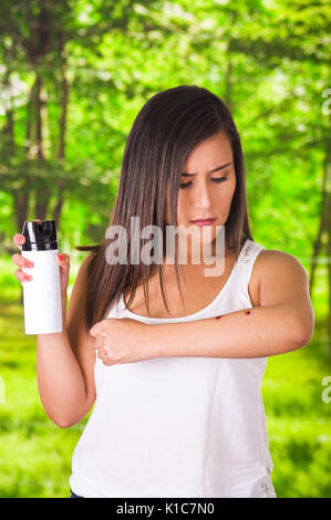 Ravvicinata di una giovane donna che soffre di prurito dopo punture di zanzara, utilizzando una spruzzatrice di oltre il morso di insetto, allergiche della pelle il concetto di trattamento, in una foresta verde sullo sfondo Foto Stock