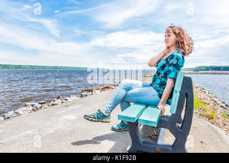 Giovane donna seduta sul banco di blu affacciato sul tranquillo fiume San Lorenzo in Portneuf, Quebec, Canada Foto Stock