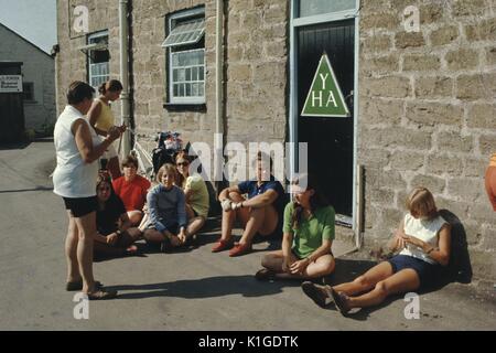 Un gruppo di ragazze adolescenti al di fuori seduta di un ostello della gioventù, una donna matura in piedi di fronte a loro e parlando al gruppo, Ostello della gioventù logo associazione visibile sulla porta dell'ostello, Inghilterra, 1966. Foto Stock