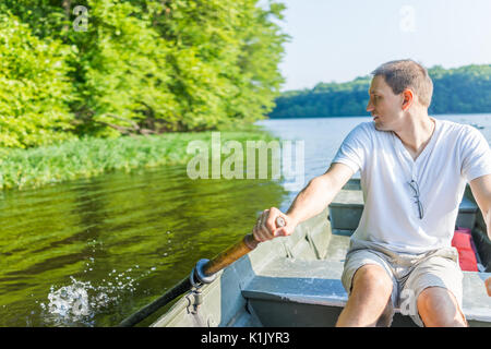 Giovane uomo barca a remi sul lago in Virginia durante l estate in camicia bianca guardando indietro agli spruzzi d'acqua Foto Stock