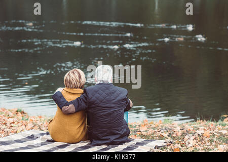 Vista posteriore della coppia matura seduta abbracciando il plaid e guardando sul lago con le anatre nel parco di autunno Foto Stock