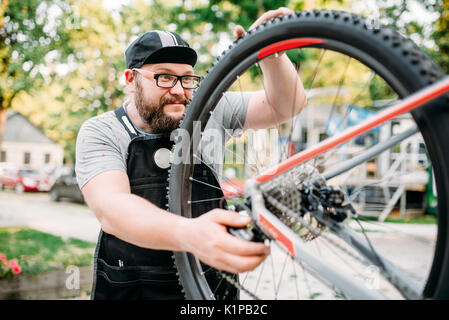 Noleggio repairman funziona con la ruota di bicicletta, ciclo workshop all'aperto. Barbuto meccanico nel grembiule Foto Stock