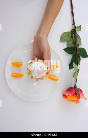 Mano che tiene la torta cesti con panna e fettine di arancia su di un piatto sul tavolo bianco e rosa. Vista dall'alto. Foto Stock