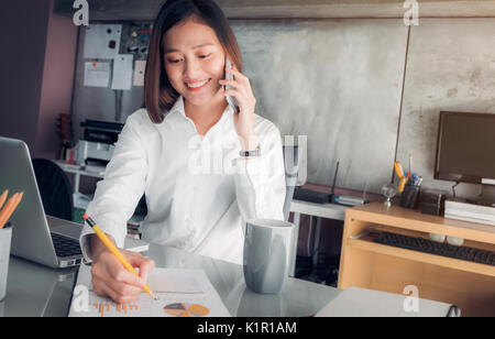 Asian imprenditrice con volto sorridente parlando al cellulare e la scrittura di analizzare i dati aziendali su foglio sulla scrivania con il collega sul telefono ,Office vita concep Foto Stock