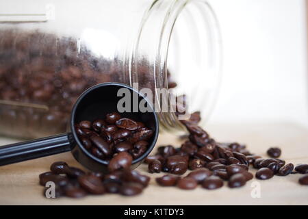 I chicchi di caffè fuoriuscito da un barattolo di vetro e un cucchiaio di misurazione su un tagliere di legno. Foto Stock