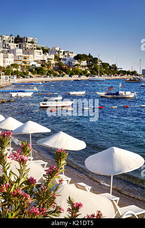 Bianchi ombrelloni e lettini vicino a laguna con barche sulla spiaggia di bodrum, Turchia Foto Stock