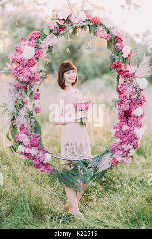 Il lato verticale della damigella sorridente con il bouquet di fiori di colore rosa e in piedi dietro il matrimonio di peonie arch situato nel soleggiato Campo. Foto Stock