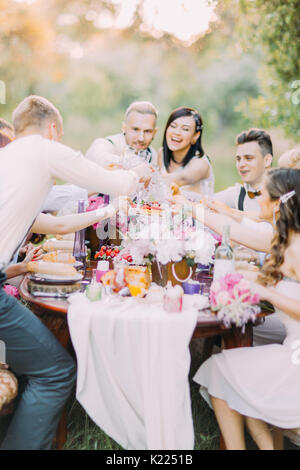 Felice di close-up verticale del delizioso sposi novelli e gli ospiti il tintinnio loro bicchieri e divertirsi presso l'impostazione tabella piena di cibo e bei fiori nella foresta. Foto Stock