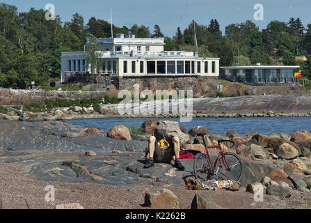 Ciclista rilassante sulla spiaggia rocciosa con grandi massi. Casa con finestre panoramiche in background Foto Stock