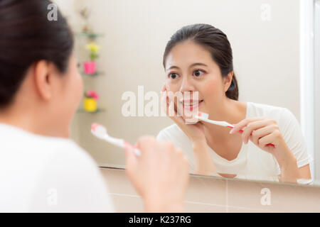 Dolce sorridente holding dello spazzolino e mettere mano sul mento cercando in corrispondenza dello specchio di riflessione la visualizzazione immagine la pelle del viso sentirsi felice in bagno a casa. Foto Stock