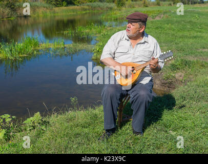 Senior man aventi il resto su un riverside seduto su un sgabello in vimini, cantare e suonare il mandolino Foto Stock