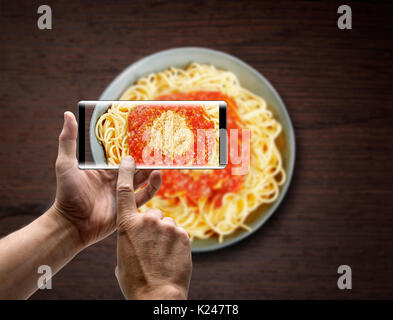 Due mani tenendo un mobile Smartphone e scattare una foto di un piatto di spaghetti alla bolognese sulla texture in legno Foto Stock