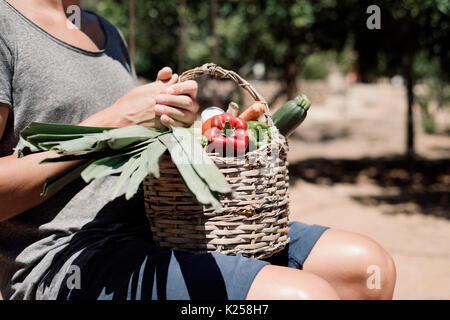 Primo piano di un giovane uomo caucasico con un rustico cesto pieno di ortaggi appena raccolti in un frutteto organico Foto Stock