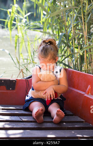 Carino bambina si siede in una barca sul lago. Ha conseguito la sua preferita il giocattolo di peluche e gode di una bella e soleggiata giornata d'estate. Infanzia, cresce la spesa Foto Stock