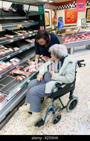 Accompagnatore o assistente aiuta una donna anziana in una sedia a rotelle scegliere un giunto di carne in un supermercato. Foto Stock