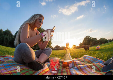 Una giovane ragazza gode di un lecca-lecca e un bicchiere di vino su un tappeto in estate mentre gli sms sul suo cellulare Foto Stock